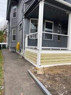 Gray house with porch, light-colored wooden skirting, white railing, and a concrete path.