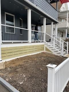 A building's porch with white railing and steps. A person is on the porch. The area below is covered in mulch.