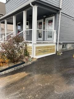 Gray house with a porch. White railing, posts, and wooden skirting. Asphalt driveway.