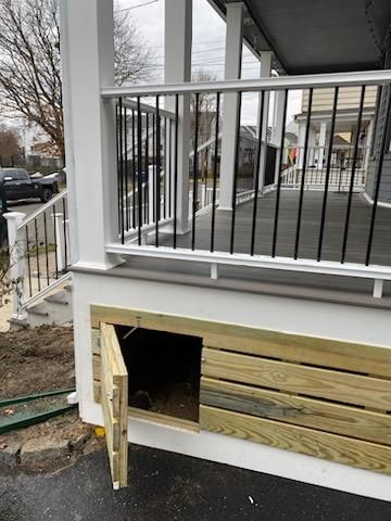 Wooden door open to a crawlspace under a porch, with black railing and white trim.