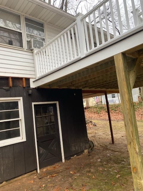 A deck with white railing overhangs a dark shed with a door and window. The structure is supported by wooden posts.