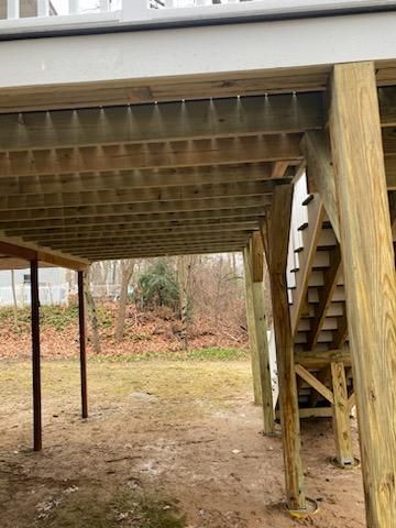 View under a wooden deck with stairs and support posts. The ground has bare grass and fallen leaves.