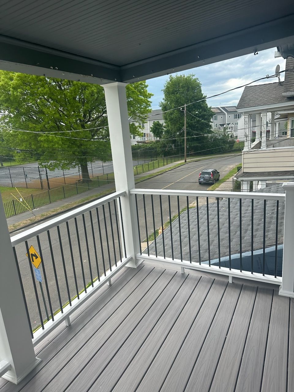 A gray porch with dark railing overlooks a street. Trees and a cloudy sky are in the background.