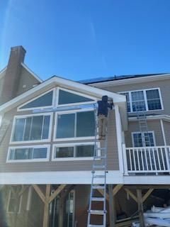 Person on ladder working on a house with large windows and a blue sky overhead.