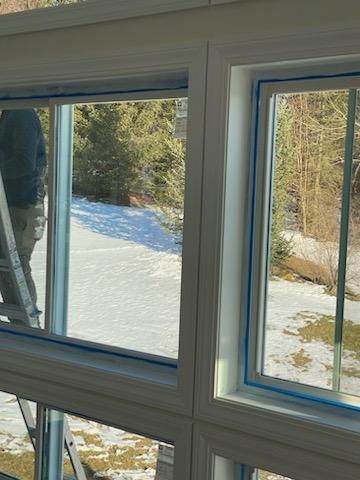 White-framed windows with blue tape, interior view of a snowy outdoor scene, person on a ladder.