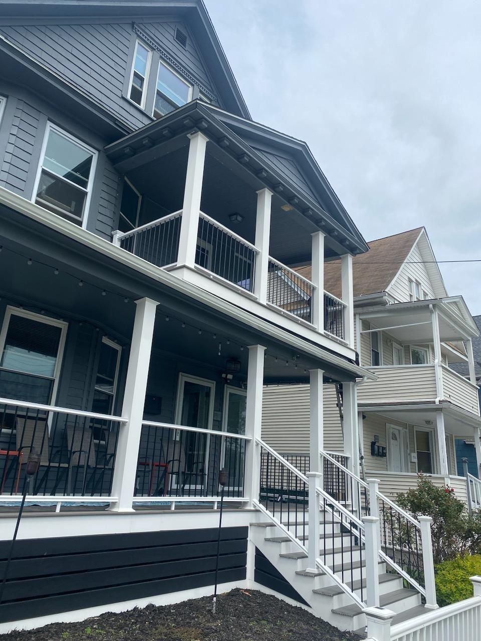 Gray and black multi-story house with porch and white railings. Cloudy sky in the background.