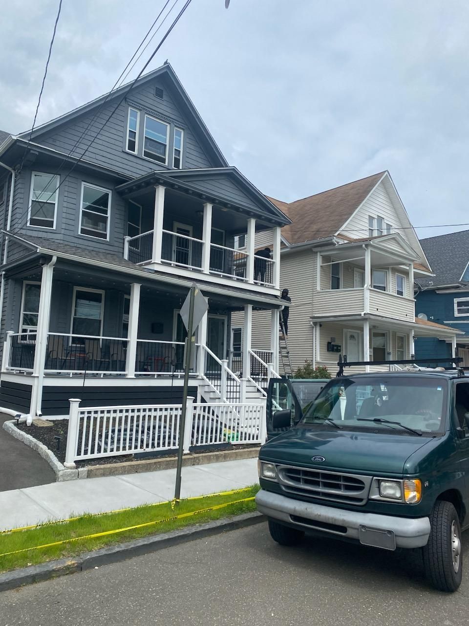 Two-story houses, one gray, one tan, with porches. A green van is parked in front of the gray house on a city street.