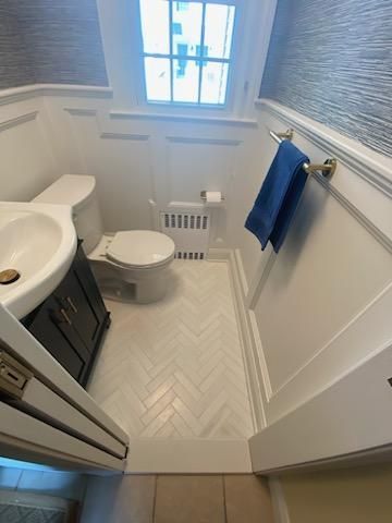 Small bathroom with white herringbone tile floor, toilet, sink, and window. Blue towel hangs on gold bar.
