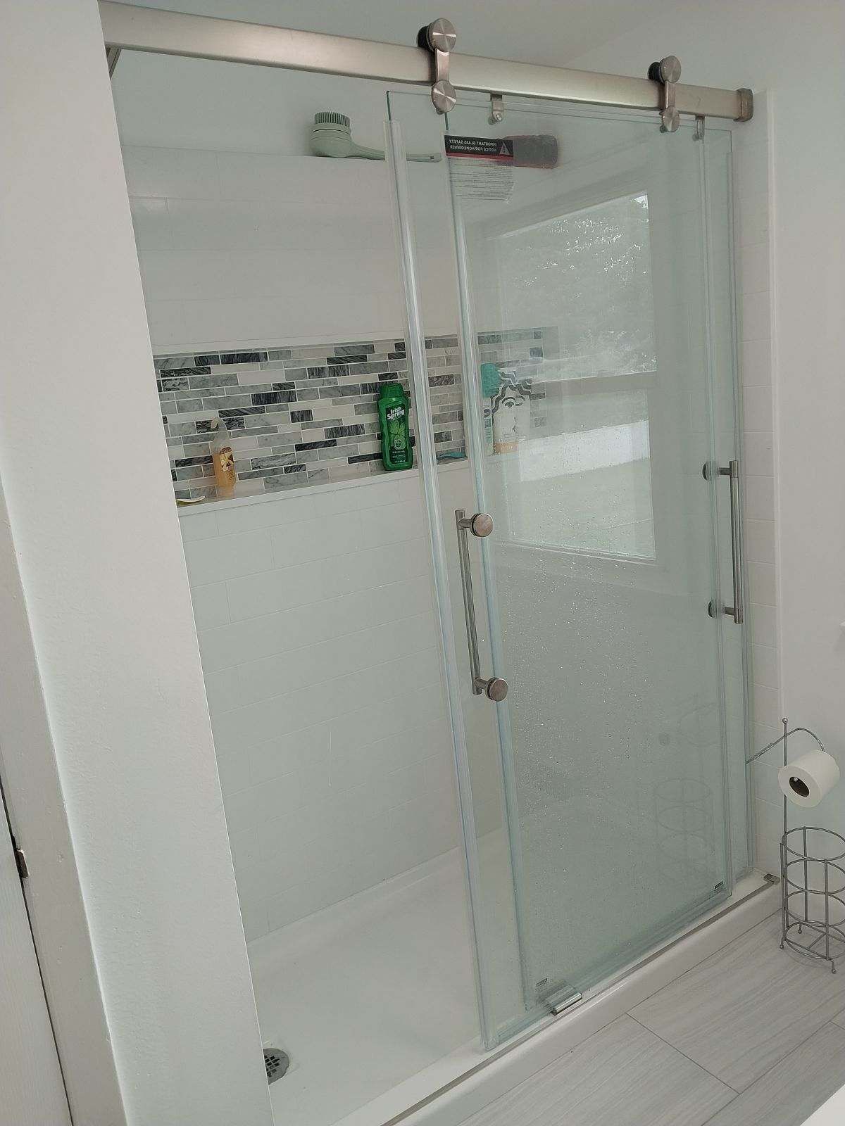 Sliding glass shower door in a white bathroom. The shower has a mosaic tile accent and a shelf with products.