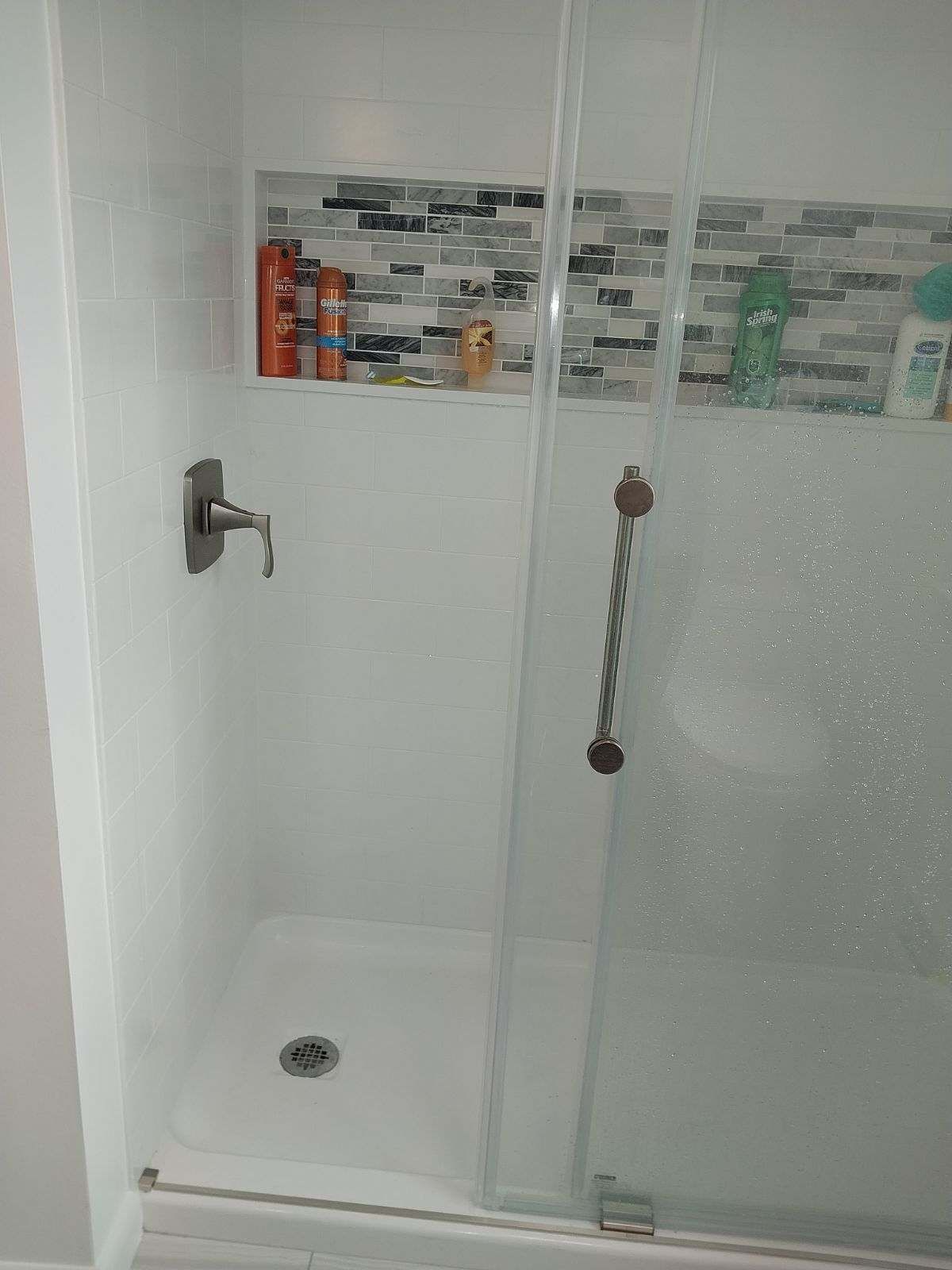 Shower stall with white walls and a glass door, a mosaic tile shelf, and toiletries.