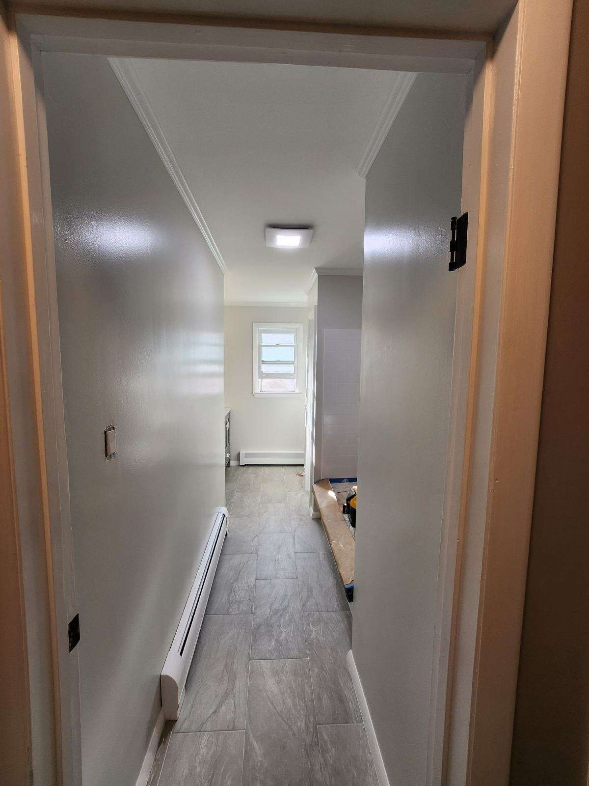 Narrow hallway with light gray walls and flooring, leading to a window.