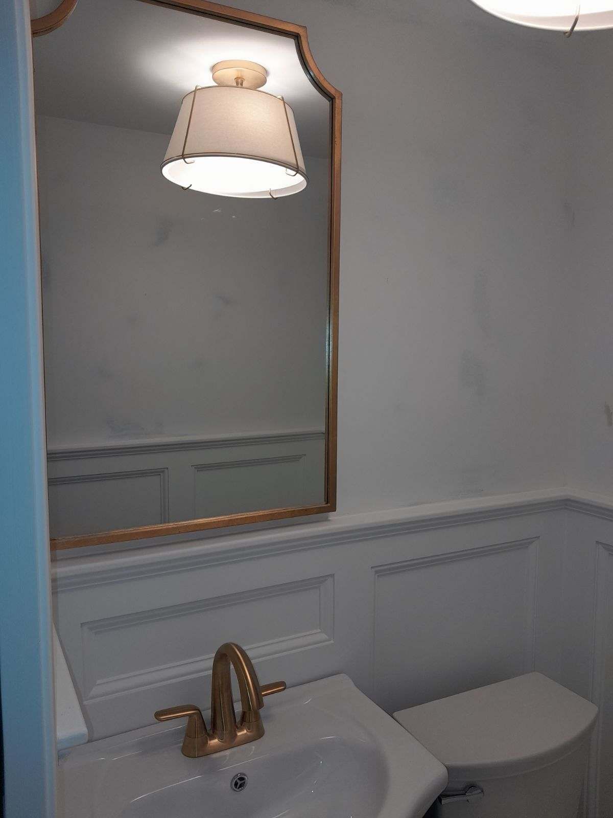 Bathroom with gold-framed mirror, white walls, sink, toilet, and lit lamp.