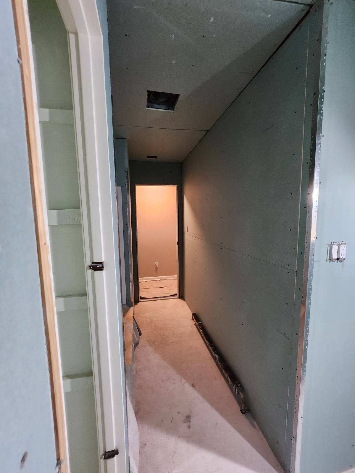 Narrow hallway under construction with drywall and unfinished floor, leading to a doorway.