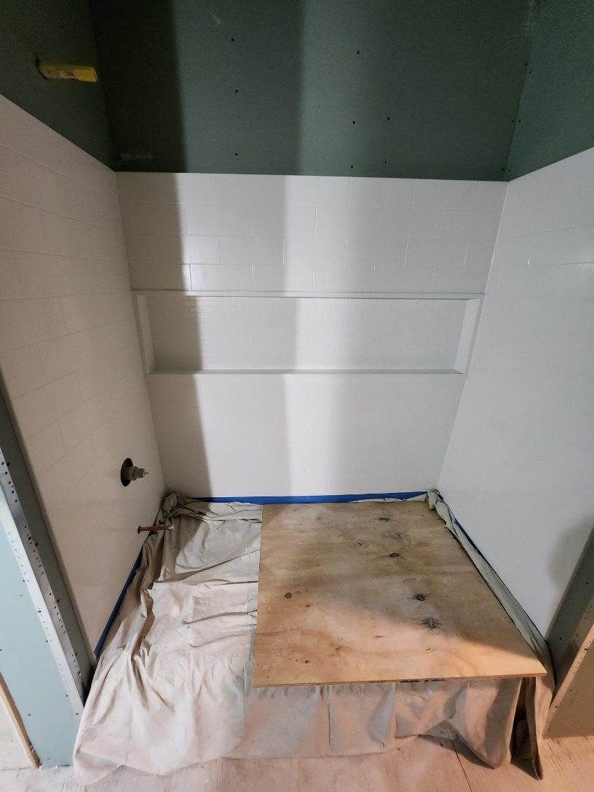 Shower stall under construction, with white walls, a shelf, and wood platform, covered in a drop cloth.