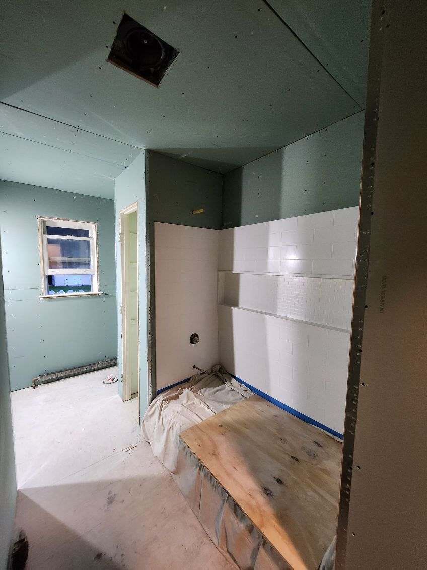 Bathroom under construction, walls of green drywall, white shower tiles, and a wooden bench.