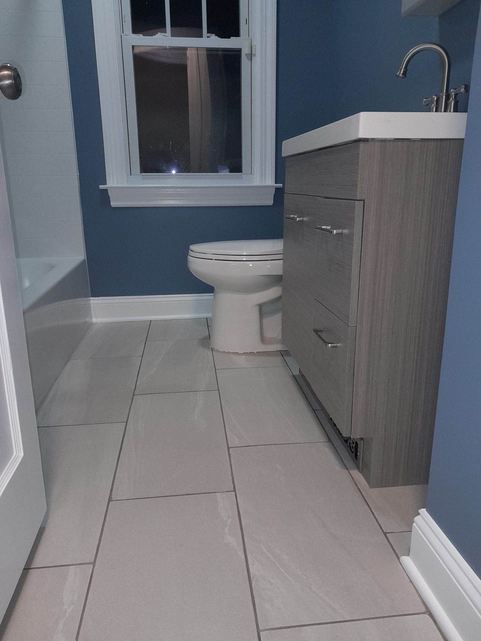 Bathroom with a gray vanity, white toilet, and blue walls. Window in the center. Gray tiled floor.