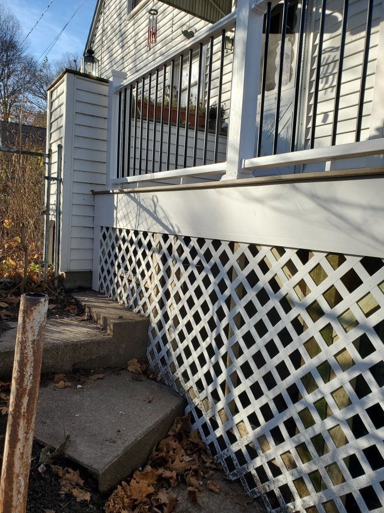 White house porch with lattice skirting and steps. Black railing, cloudy sky.