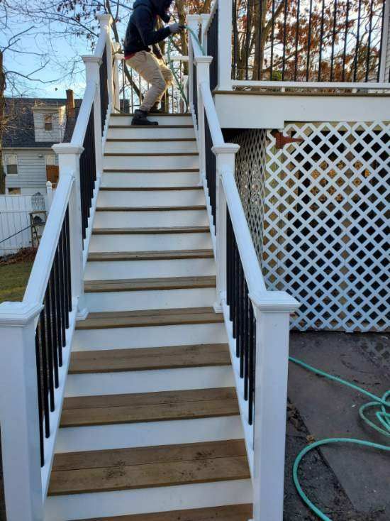 Person on outdoor stairs, white risers, wooden treads, black railing. Deck in background, green hose to the right.