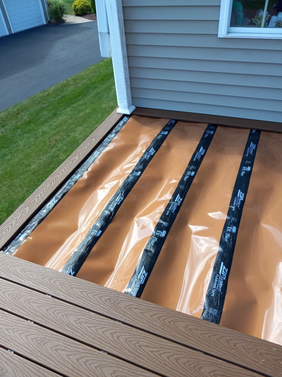 Deck underlayment with brown boards, black tape, and orange membrane next to a house with beige siding.