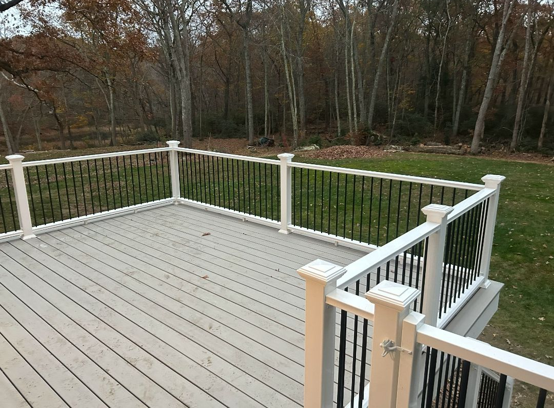 Deck with white posts, black railings, and gray floorboards. Backyard with trees in background.
