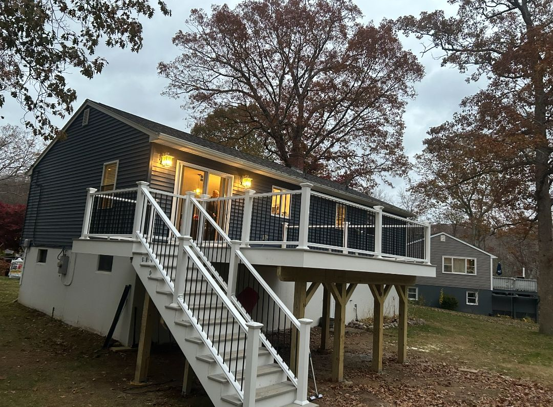 A raised house with a white deck and staircase. Dark siding, windows with warm light, and surrounding trees.