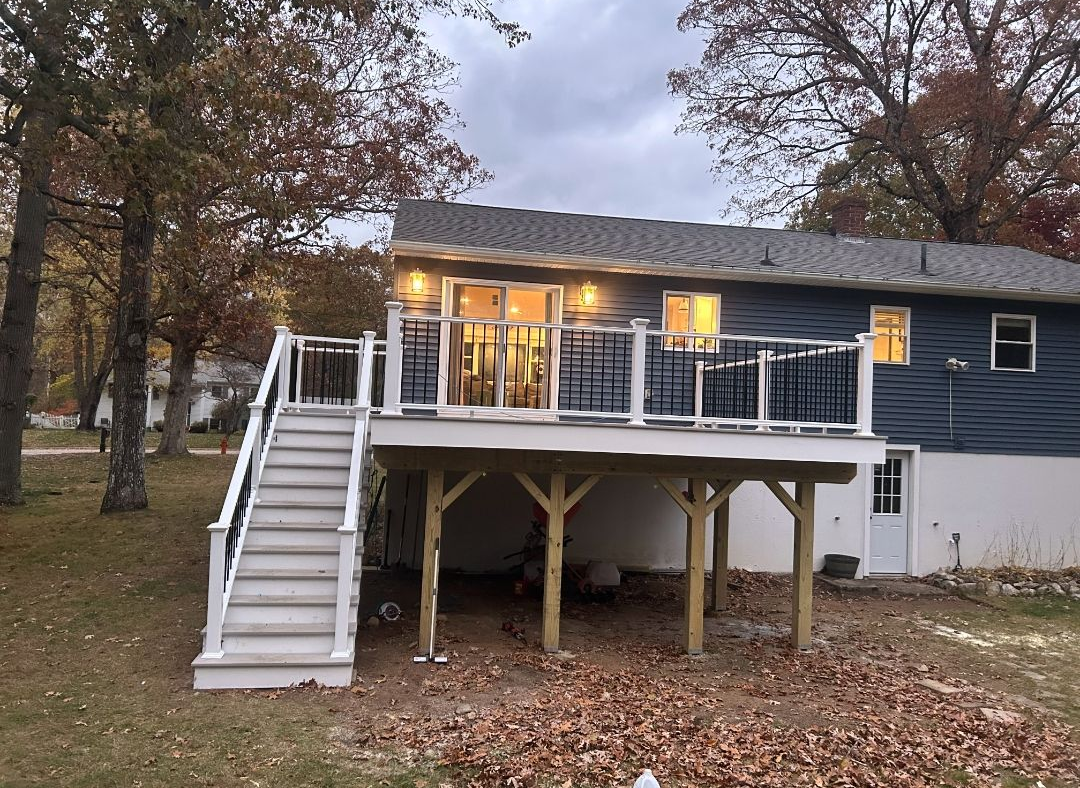 Back of a house with a deck. Deck has stairs and white railing. Blue siding, fall leaves on the ground.