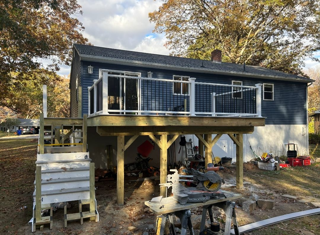 Newly built deck with a dark blue house. White and black railing on the deck with tools and wood visible below.