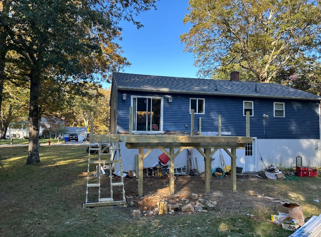 A deck under construction at a blue-sided house. Wooden framing, stairs, and tools are visible in the yard.