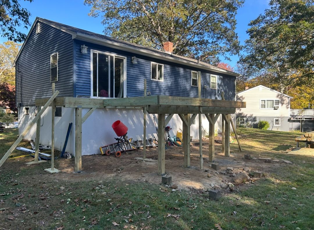 Wooden deck being built beneath a two-story blue house. Construction materials and tools visible.