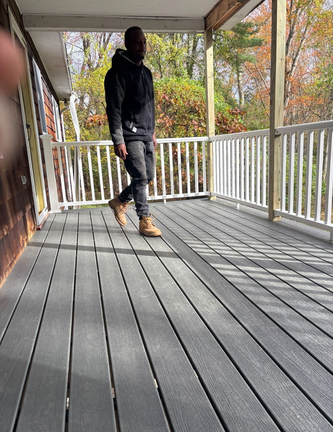 Person on a gray deck wearing a black hoodie and jeans, standing near a white railing. Trees in the background.