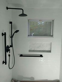 Black shower fixtures in a white tiled shower with window and shelf, in a bathroom.