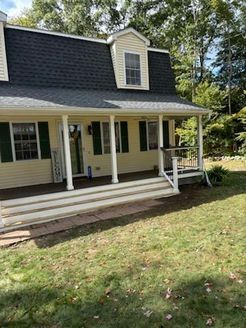 Yellow house with a black roof and porch. Green shutters and a lawn.