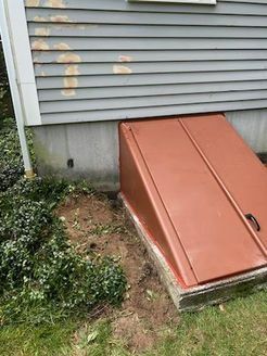 Red cellar door with angled top against a gray house with damaged siding.