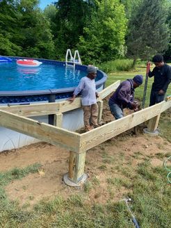 Three people constructing a wooden deck around an above-ground pool; sunny outdoor setting.