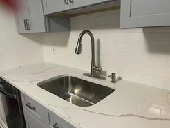 Stainless steel sink with faucet on white countertop, under grey cabinets, white tile backsplash.