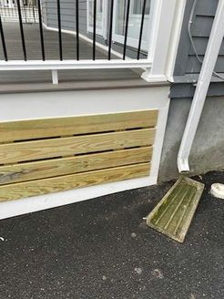 Wooden slatted paneling below a white porch with black railing. Gray siding and a downspout are visible.