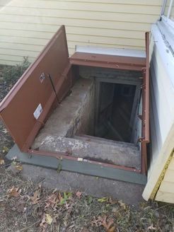 Brown metal cellar door open, leading to concrete steps. Located beside a light yellow house.