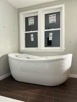 White bathtub wrapped in plastic under a window in a bathroom with dark brown tile floor.