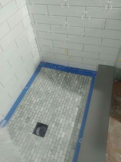 A shower under construction with white brick-style wall tiles and a mosaic tile floor, drain, and a blue trim.
