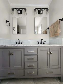 Gray bathroom vanity with two sinks, mirrors, and sconces. Beige towel, dark hardware, and marble countertop.