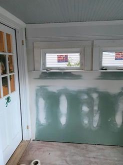 Interior room under renovation, drywall with visible joint compound below two small windows, door on the left.
