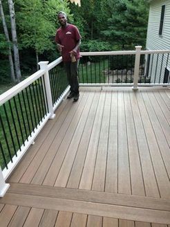 Man stands on a wooden deck with white railing and black spindles, holding a beverage, trees in the background.