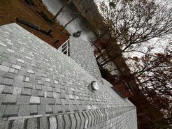 Gray shingle roof of a house with a chimney and vent against a backdrop of trees and a cloudy sky.
