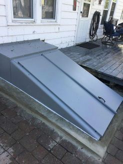 Gray metal cellar door on a concrete base, leading up to a white house with a porch and brick patio.