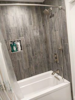 Bathroom with gray wood-look tile, white tub, silver shower fixtures, and built-in shelf with toiletries.