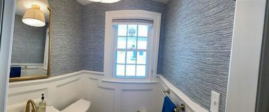 Bathroom with blue textured wallpaper, white trim, gold mirror, and window.
