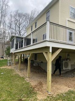 A two-story house with a wooden deck and supports. A person stands underneath the deck, near AC units.