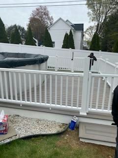 A white deck with a pool and fence. Brown grass and a white house in the background. Overcast sky.