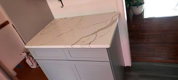 A cabinet with a light, marbled countertop. Gray cabinets and wooden floor are visible. A plant is in the window.