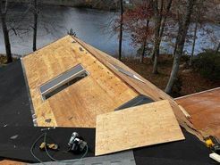 Roof with exposed plywood, skylights, and tools; near a body of water.
